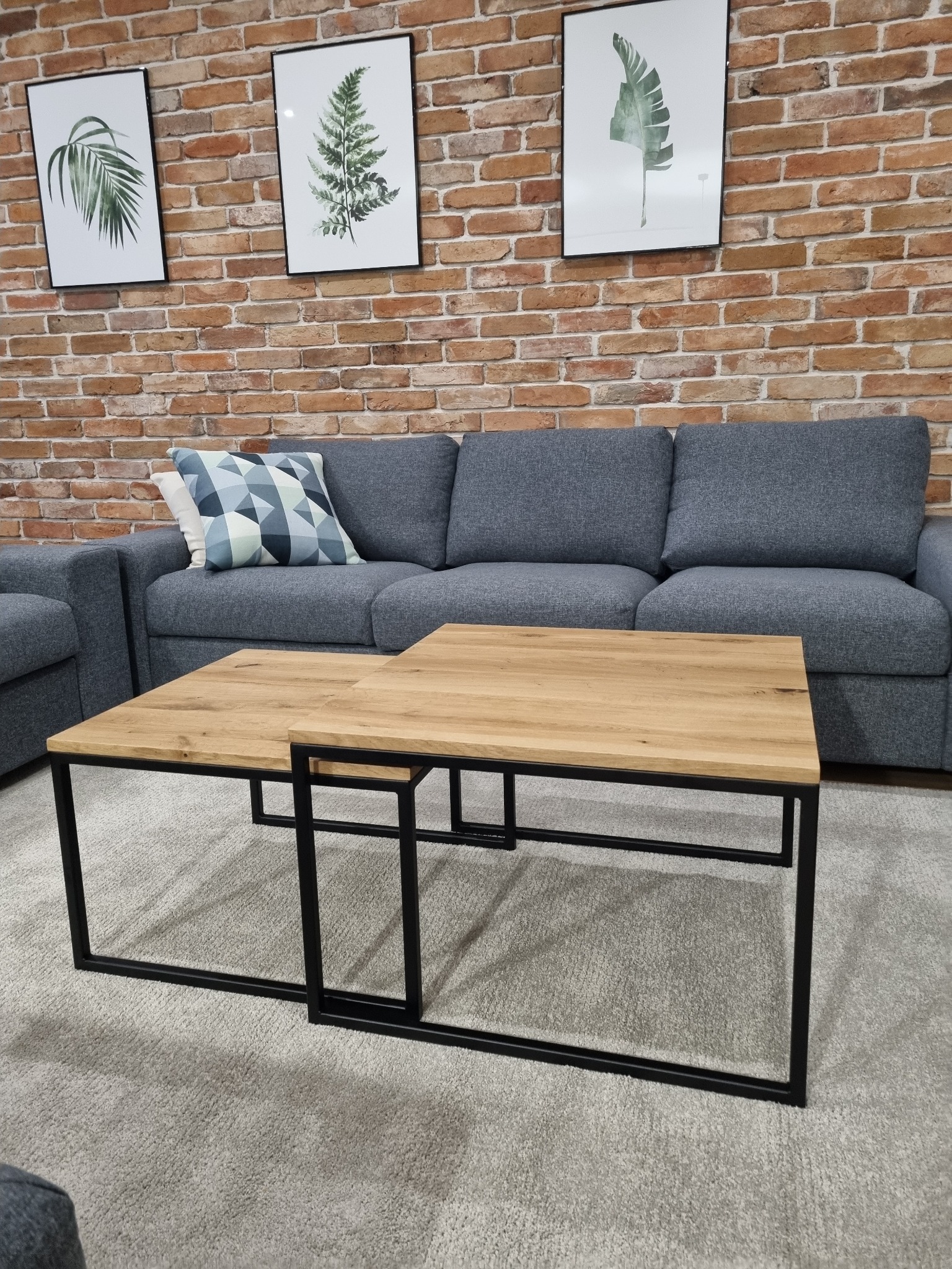 Dwa stoliki kawowe z drewnianymi blatami i czarnymi metalowymi nogami, ustawione na szarym dywanie, na tle ceglanej ściany z trzema obrazami roślinnymi.