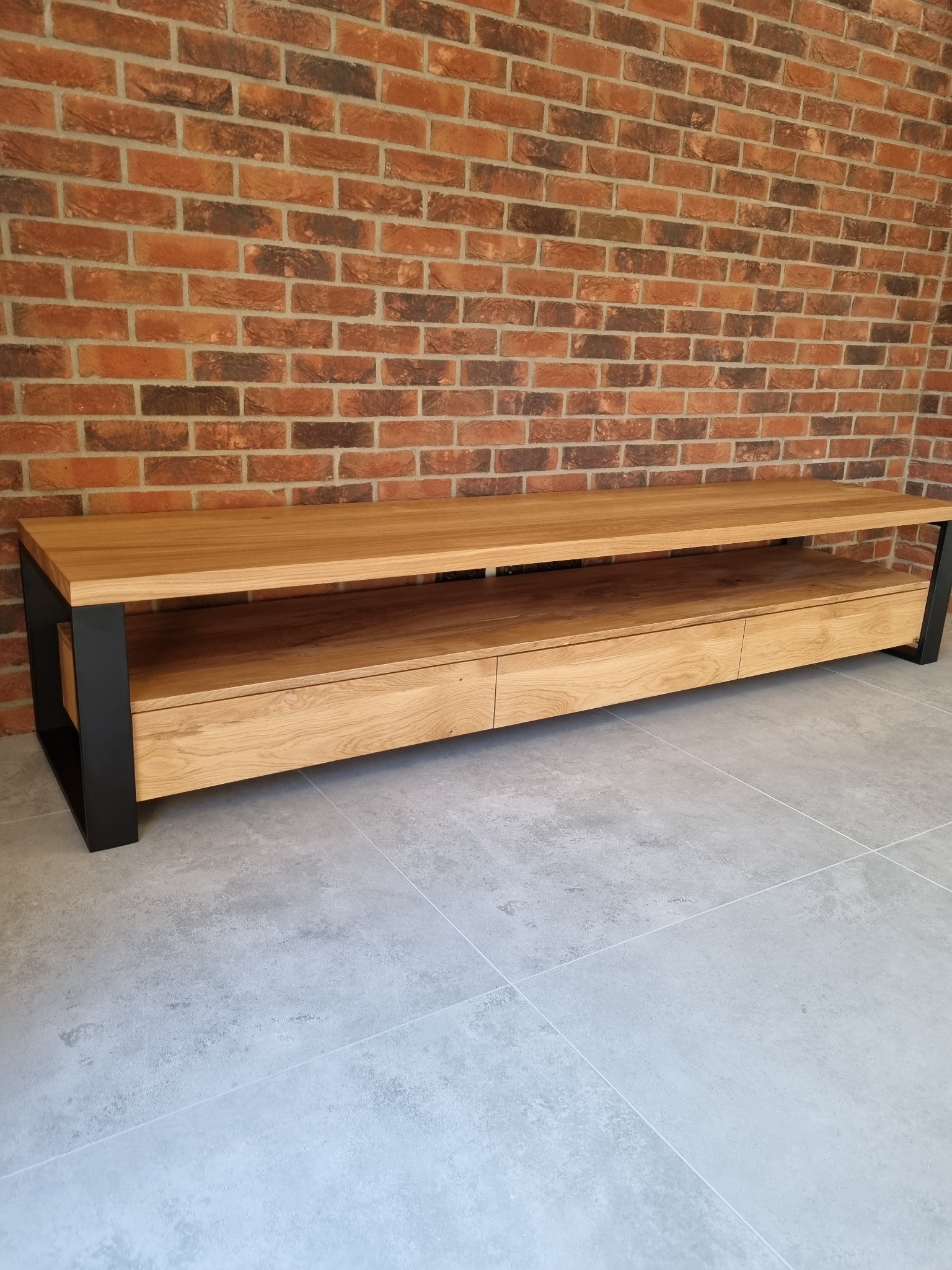 Drewniana szafka RTV z trzema szufladami i czarnymi metalowymi nogami, ustawiona na szarych płytkach pod ceglaną ścianą.