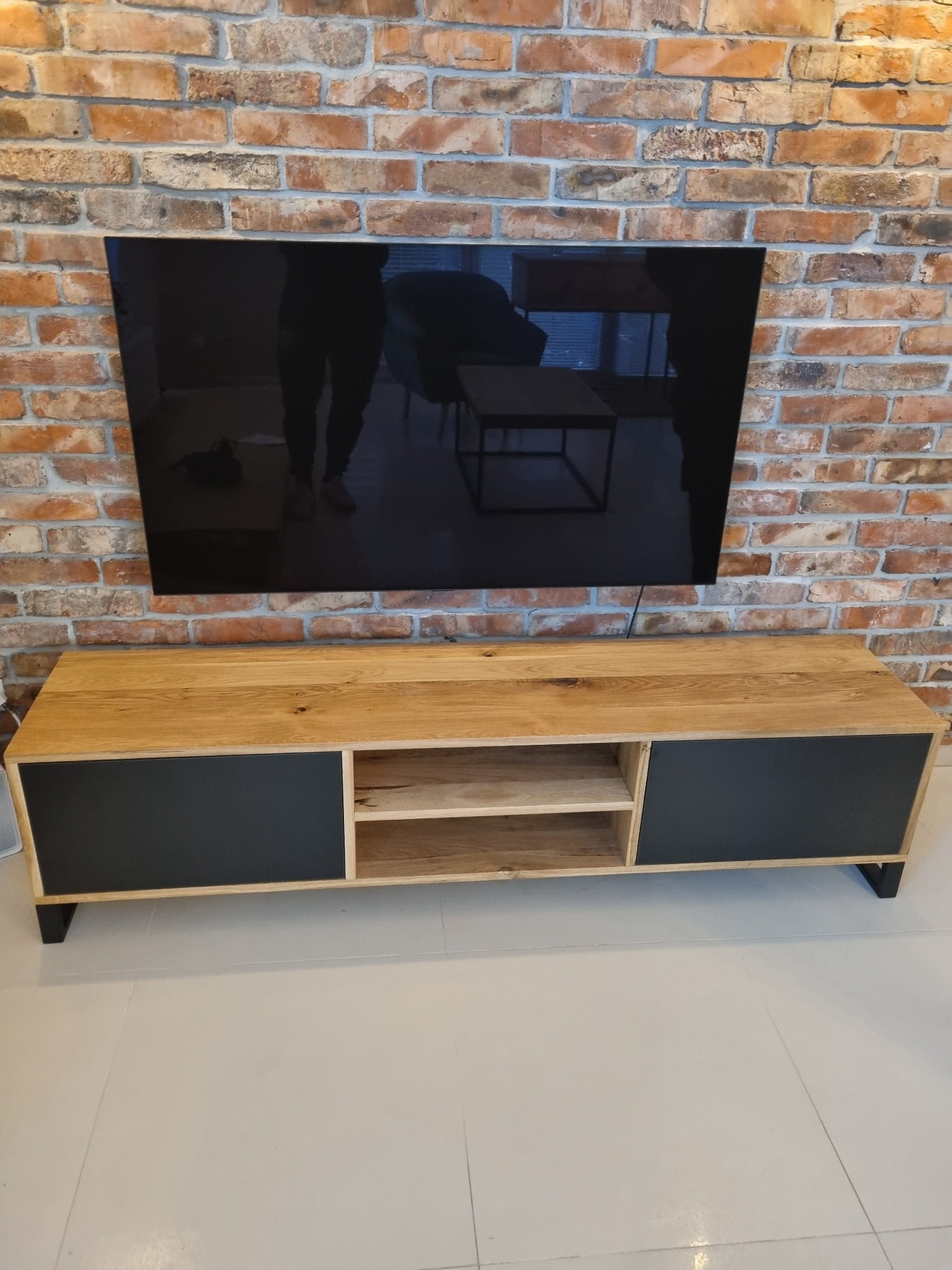 Drewniana szafka RTV z czarnymi frontami, wykonana na wymiar w Opalenicy, ustawiona pod telewizorem na tle ściany z czerwonej cegły.