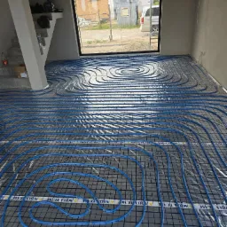 Niebieskie rury ogrzewania podłogowego ułożone na szarej macie w jasnym pomieszczeniu z dużym oknem. Widoczne elementy konstrukcyjne budynku.