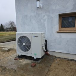 Instalmax Piotr Szachnowski - Biała pompa ciepła YORK na betonowej podstawie przy ścianie budynku z widocznymi przewodami i kostką brukową. Tło: drzewa, pochmurne niebo.