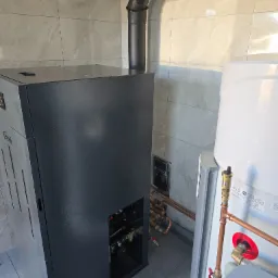 Szary piec CO marki Ferroli z czarnym kominem i biały zasobnik ciepłej wody użytkowej Atlantic z miedzianymi rurami w kotłowni wykończonej płytkami.