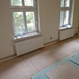 Pokój w trakcie remontu z dwoma białymi grzejnikami pod oknami, widoczne rury instalacji grzewczej oraz butelka wody na parapecie.