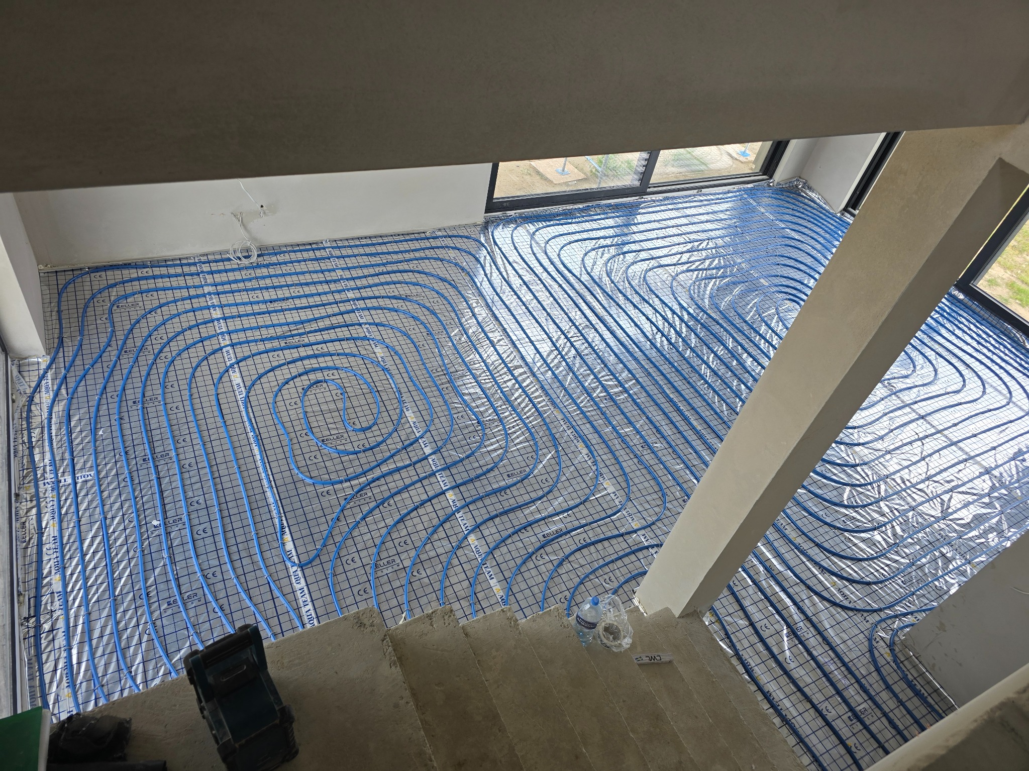 Niebieskie rury ogrzewania podłogowego ułożone na folii z nadrukiem, widok z góry, surowy stan wykończenia wnętrza, okna w tle.