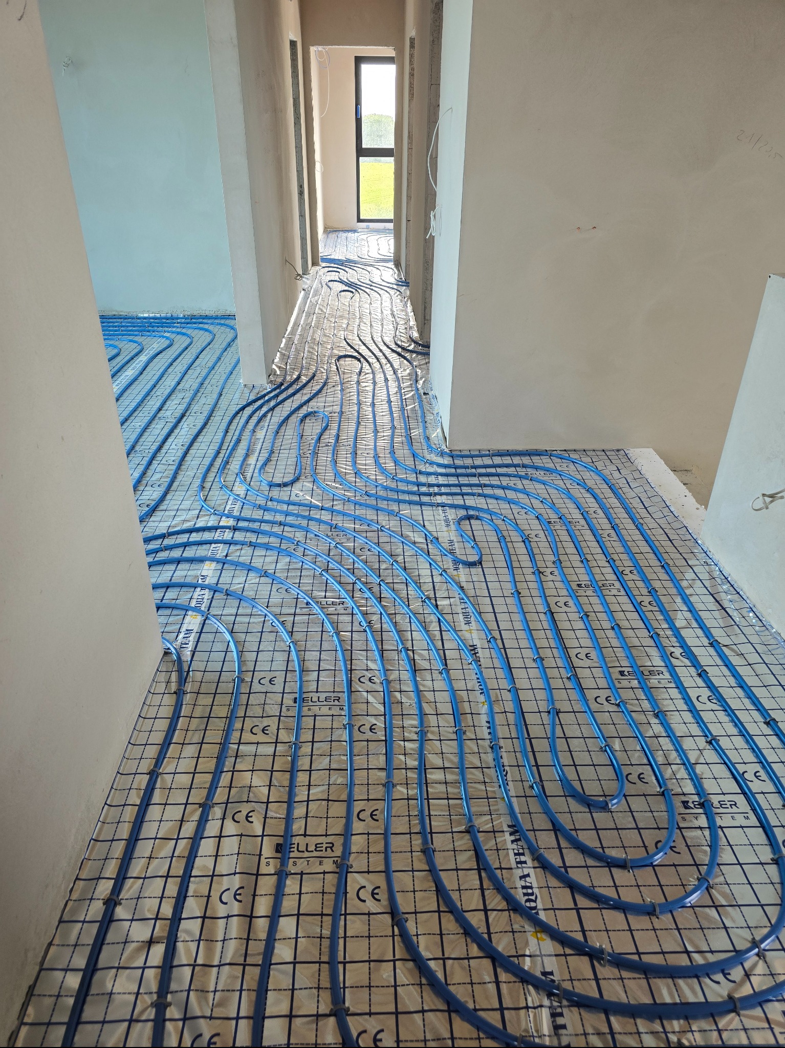 Niebieskie rury ogrzewania podłogowego ułożone na folii z rastrem w surowym wnętrzu. Widok z perspektywy podłogi, w tle widoczne kolejne pomieszczenia.