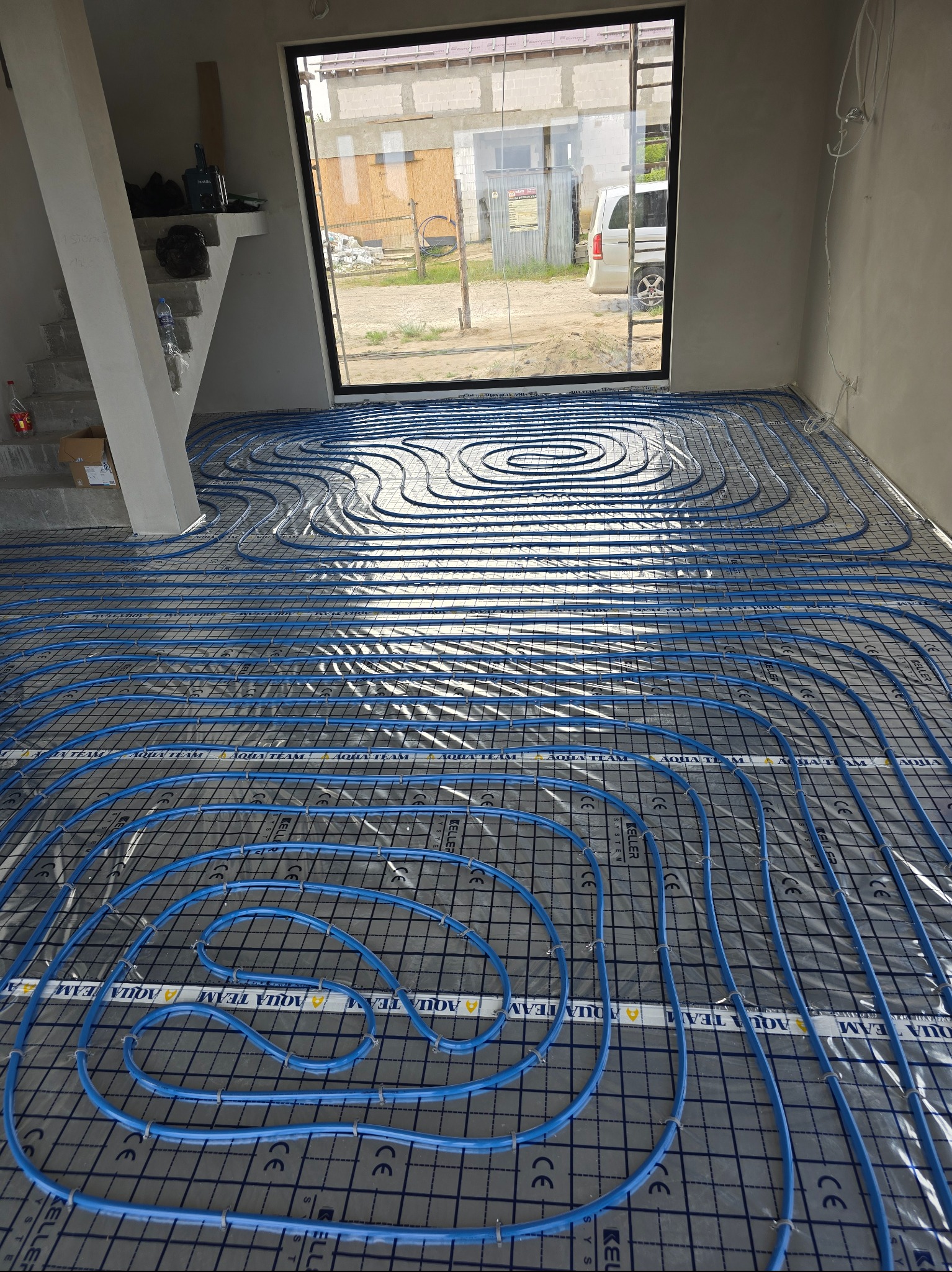 Niebieskie rury ogrzewania podłogowego ułożone na szarej macie w jasnym pomieszczeniu z dużym oknem. Widoczne elementy konstrukcyjne budynku.