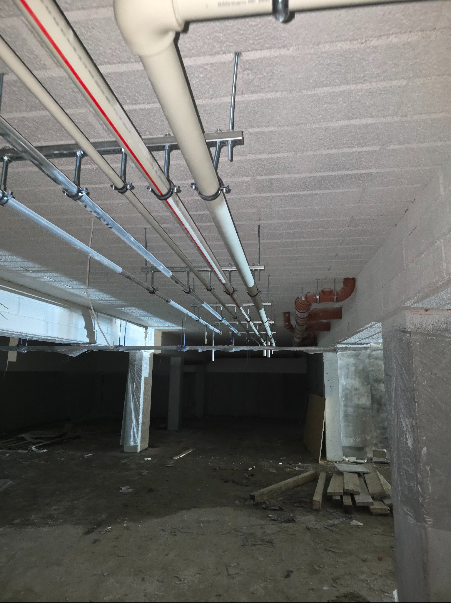 Instalacja rur pod stropem w surowym wnętrzu. Białe i szare rury z czerwonym paskiem, mocowane metalowymi uchwytami. Widoczne elementy instalacji kanalizacyjnej.