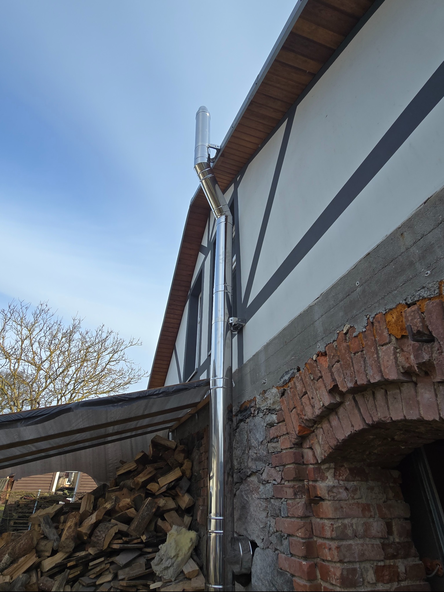Błyszczący, stalowy komin zewnętrzny zamontowany przy ścianie domu z muru pruskiego, z widocznym składem drewna opałowego pod zadaszeniem.