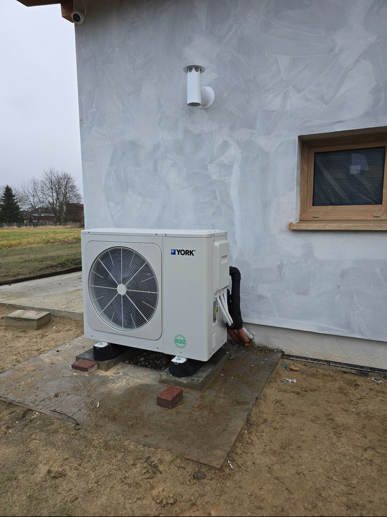 Biała pompa ciepła YORK na betonowej podstawie przy ścianie budynku z widocznymi przewodami i kostką brukową. Tło: drzewa, pochmurne niebo.