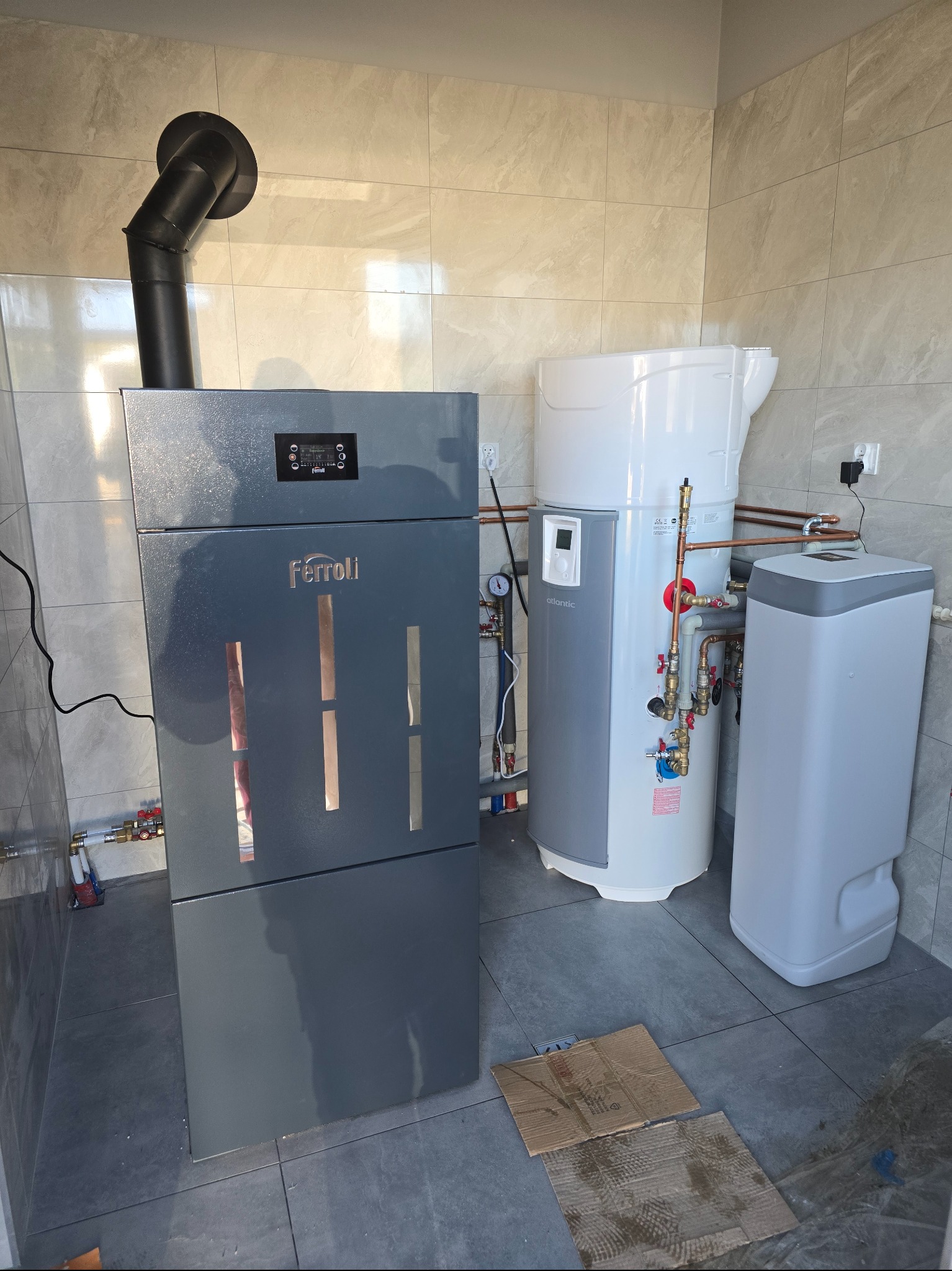 Nowoczesna kotłownia z piecem Ferroli, zasobnikiem Atlantic i zmiękczaczem wody. Instalacja w pomieszczeniu wykończonym płytkami w odcieniach szarości.