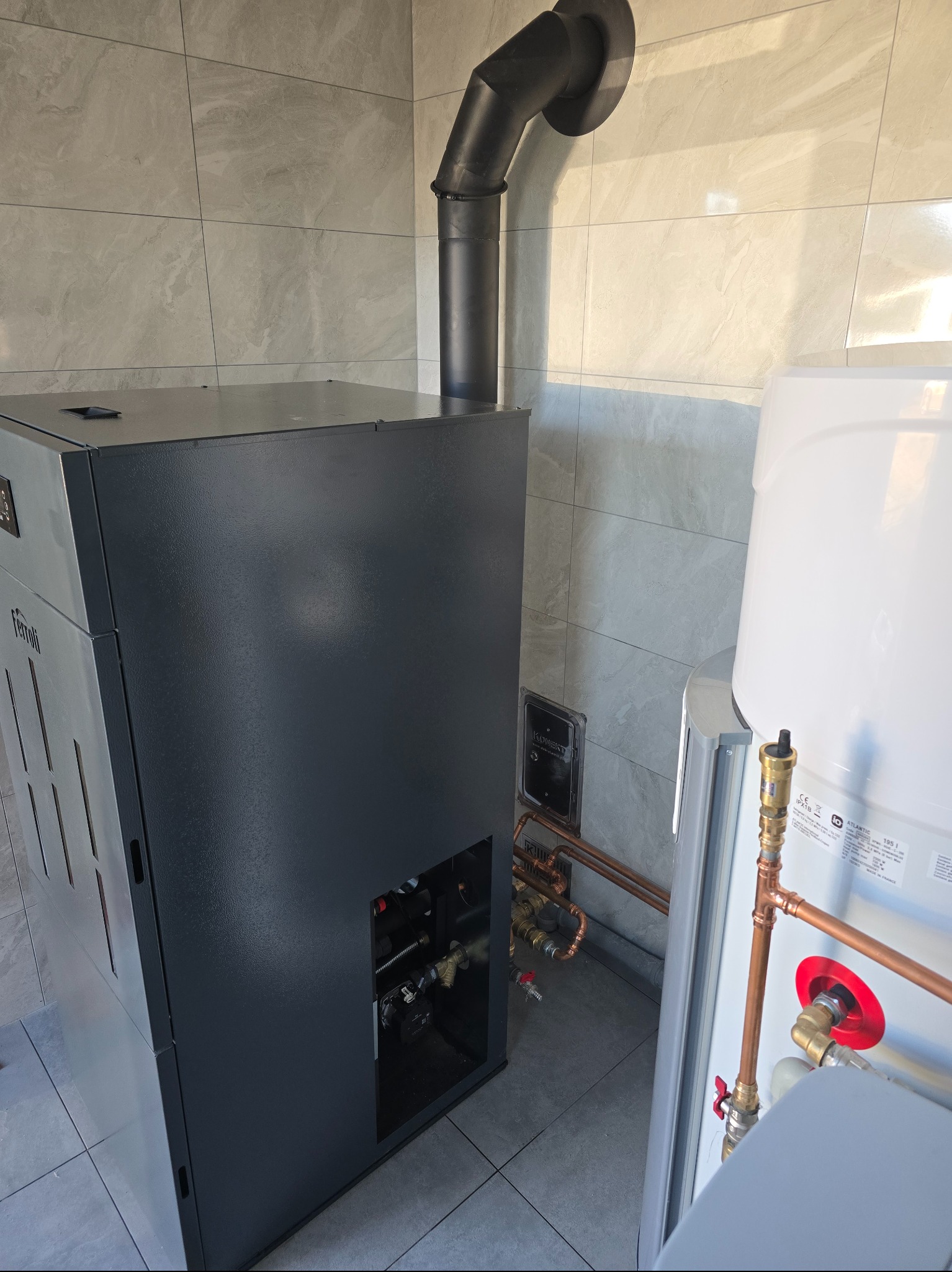 Szary piec CO marki Ferroli z czarnym kominem i biały zasobnik ciepłej wody użytkowej Atlantic z miedzianymi rurami w kotłowni wykończonej płytkami.