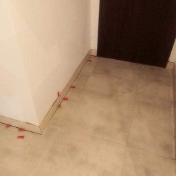 Bad Bud - Ułożone, beżowe płytki podłogowe w rogu pomieszczenia, z czerwonymi krzyżykami dystansowymi przy ścianie i progu drzwi.