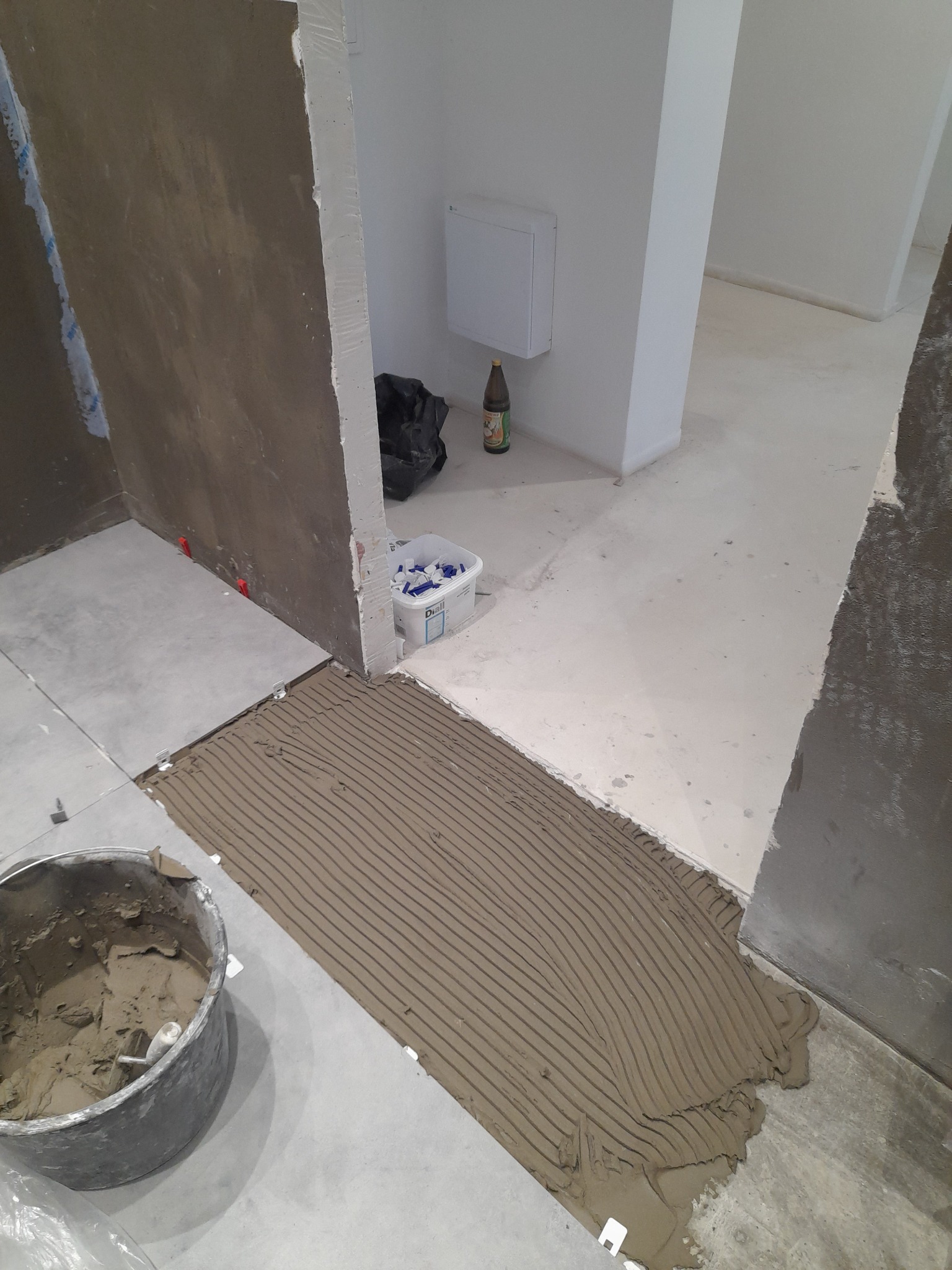 Przygotowanie do ułożenia płytek podłogowych: warstwa kleju z charakterystycznymi rowkami, wiadro z zaprawą, krzyżyki dystansowe i poziomice w trakcie prac wykończeniowych w pomieszczeniu.