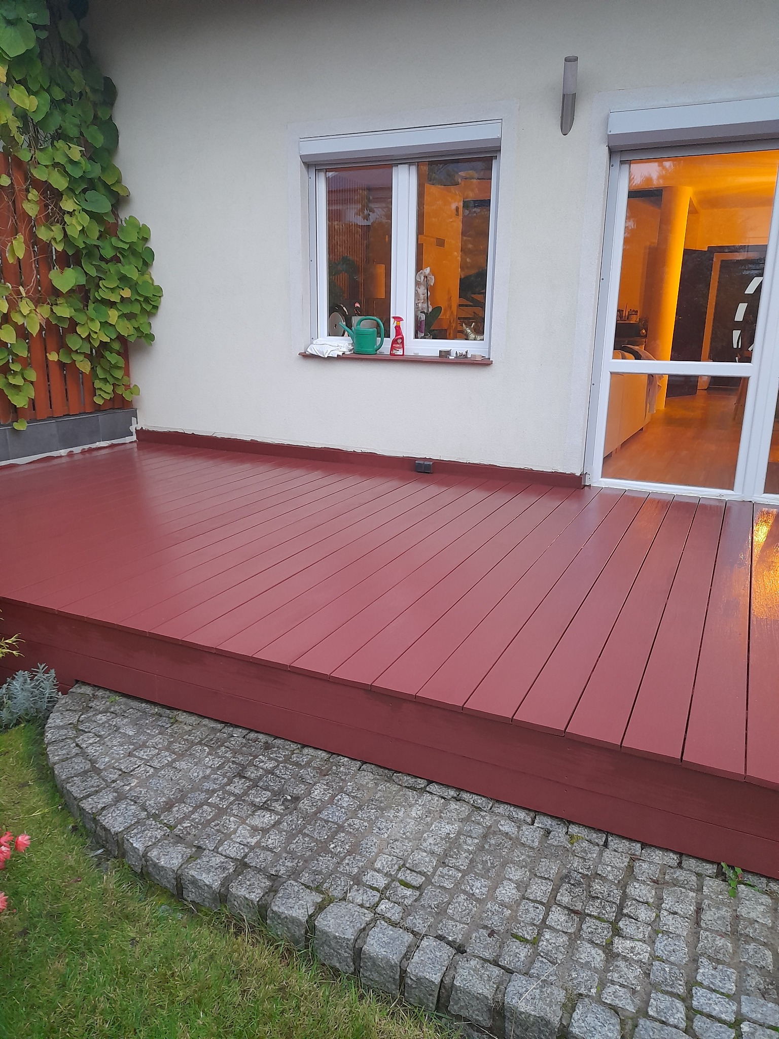 Czerwony, drewniany taras przylegający do białego budynku z oknem, na którym stoi zielona konewka i czerwona butelka, obok drzwi balkonowych z widocznym wnętrzem, wykończony kostką granitową...