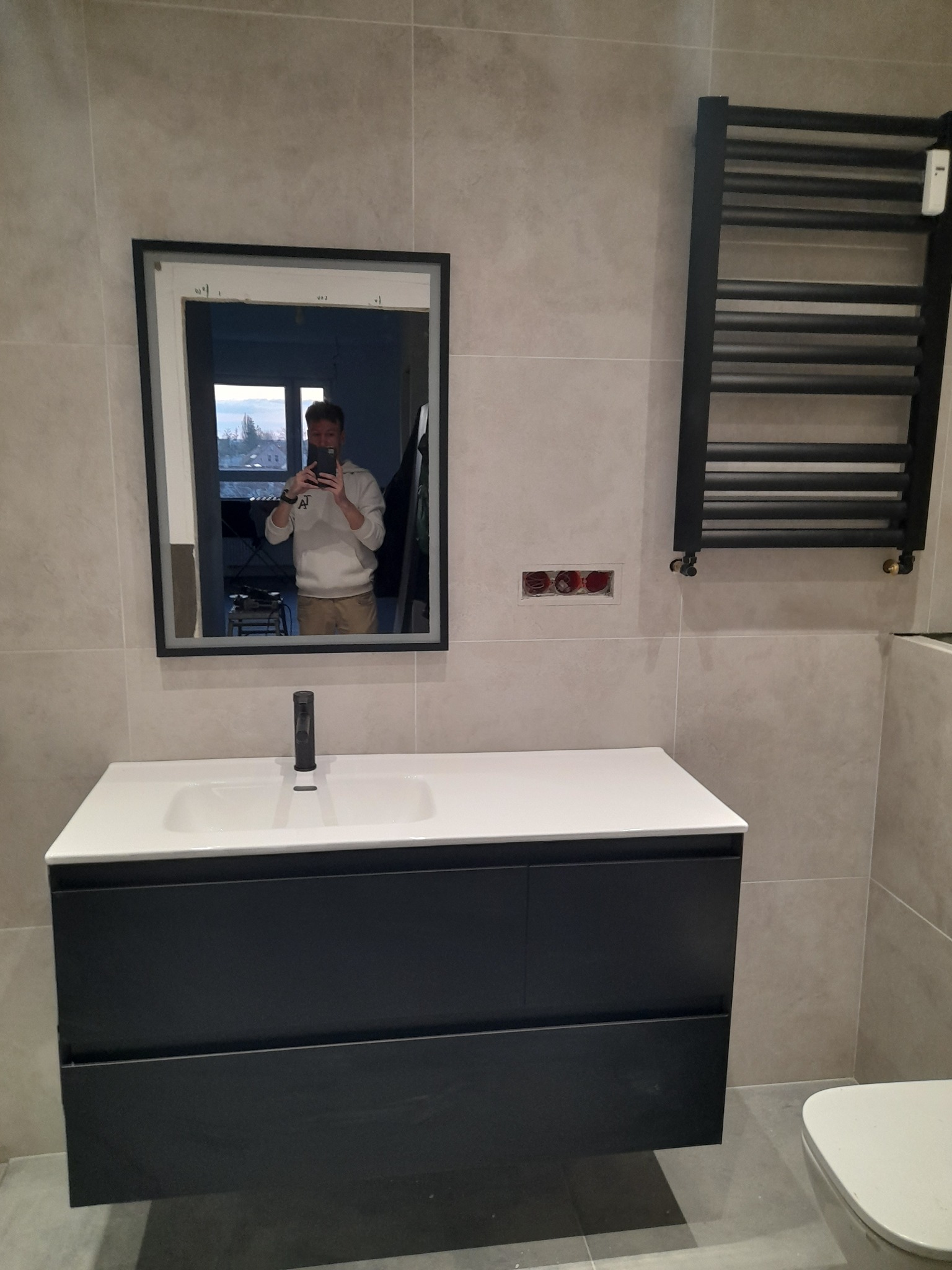 Nowoczesna łazienka z umywalką nablatową, czarną szafką pod umywalką, grzejnikiem drabinkowym i lustrem, w którym odbija się osoba robiąca zdjęcie.