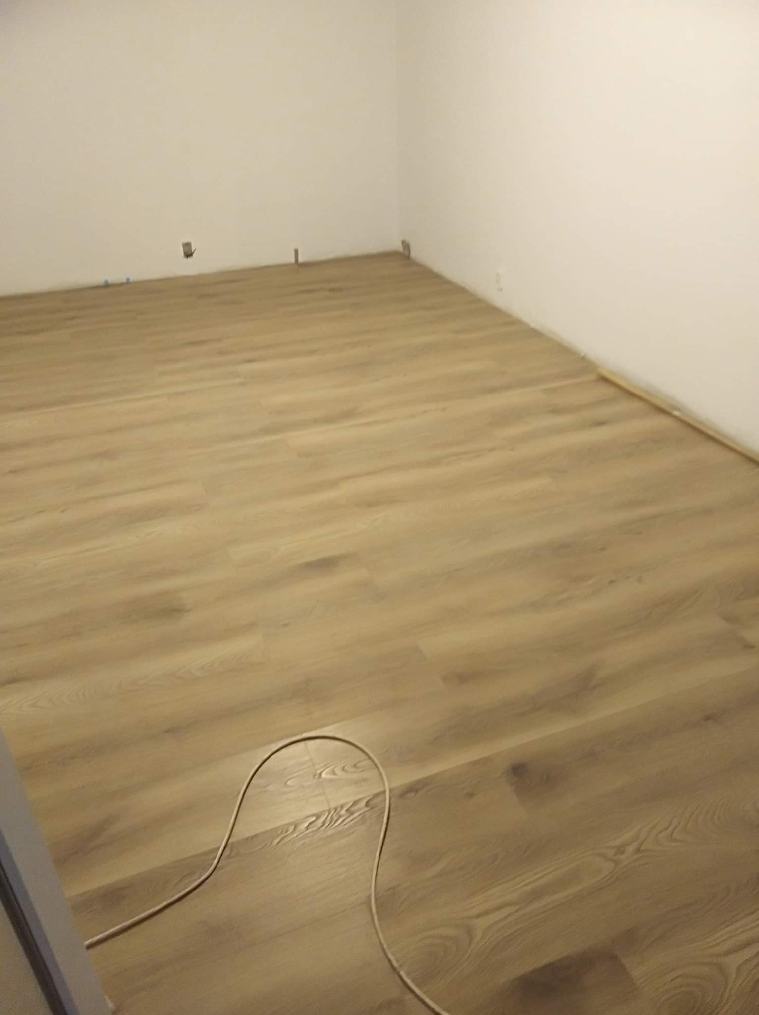 Jasny, drewnopodobny parkiet ułożony w pustym pokoju z białymi ścianami; widoczny kabel w rogu.