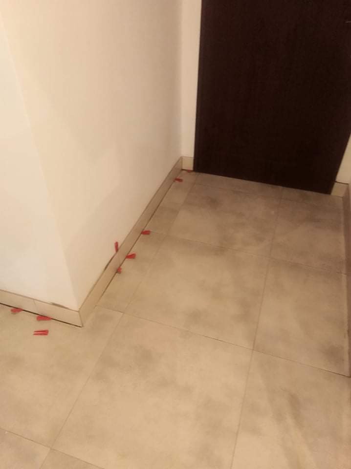 Ułożone, beżowe płytki podłogowe w rogu pomieszczenia, z czerwonymi krzyżykami dystansowymi przy ścianie i progu drzwi.