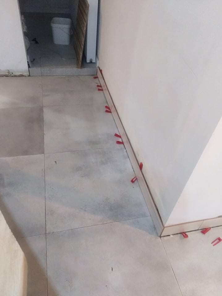 Ułożone szare płytki podłogowe z czerwonymi klipsami dystansowymi przy ścianie, widok z góry, w tle białe wiadro.