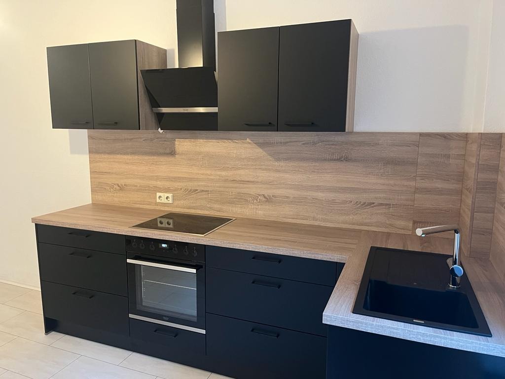 Nowoczesna, czarna zabudowa kuchenna z drewnopodobnym blatem i panelem ściennym, widok na płytę indukcyjną i zlewozmywak.