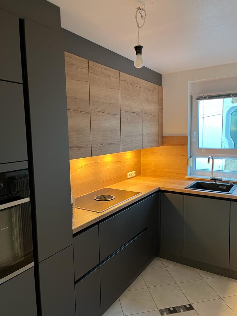 Nowoczesna kuchnia z szarymi matowymi szafkami dolnymi i drewnopodobnymi szafkami górnymi, podświetlona listwą LED, z czarnym zlewem i płytą indukcyjną.