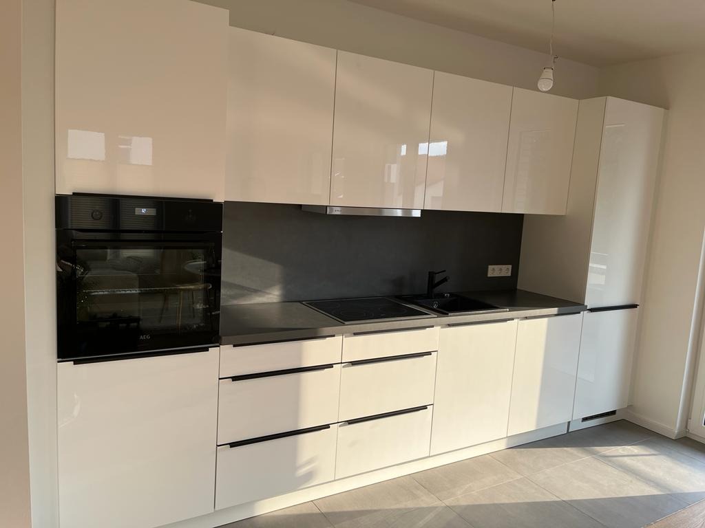 Nowoczesna, minimalistyczna kuchnia z białymi, lakierowanymi frontami, czarnym blatem i sprzętem AGD w zabudowie; widoczne subtelne, poziome uchwyty i szara ściana nad blatem.