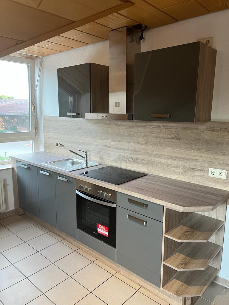 Szara zabudowa kuchenna z drewnopodobnym blatem i otwartymi półkami, widok na płytę indukcyjną i zlewozmywak, jasne płytki na podłodze.