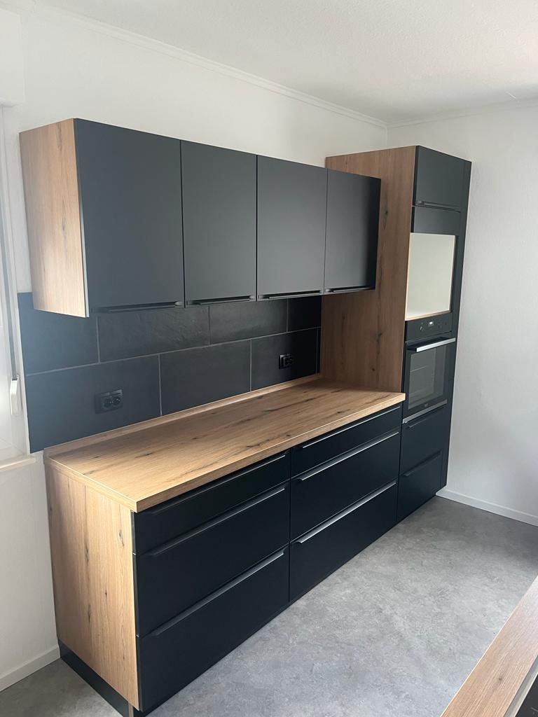 Nowoczesna kuchnia z czarnymi szafkami i drewnianym blatem, czarne płytki na ścianie, szare panele podłogowe, zabudowa piekarnika z drewnianymi bokami.