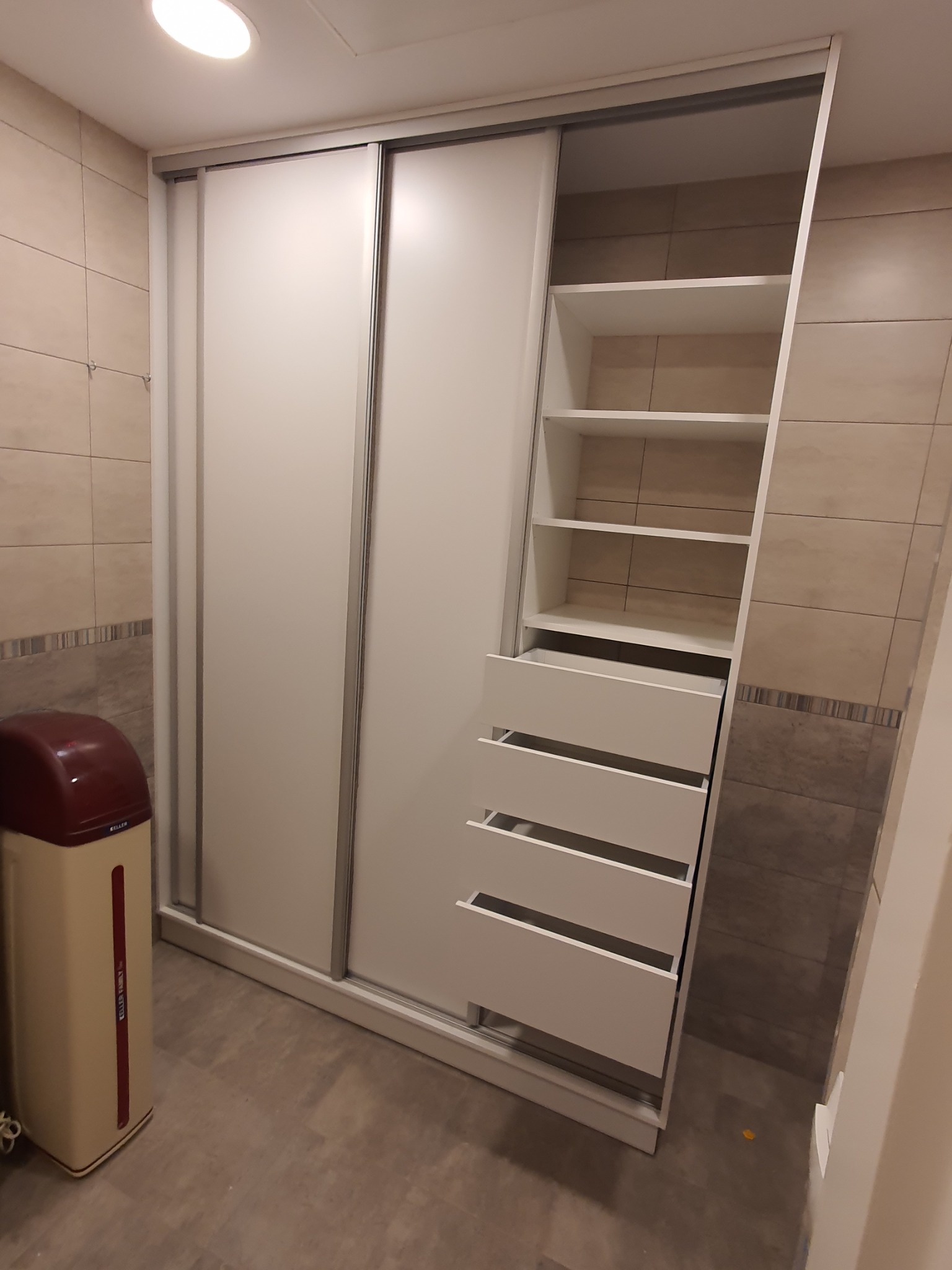 Biała szafa przesuwna z otwartą częścią z półkami i szufladami, widoczny fragment łazienki z brązowymi płytkami i zmiękczacz wody Keller.