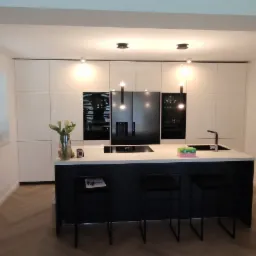 Nowoczesna kuchnia z białymi szafkami i czarną wyspą, oświetlona wiszącymi lampami, z drewnianą podłogą ułożoną w jodełkę.