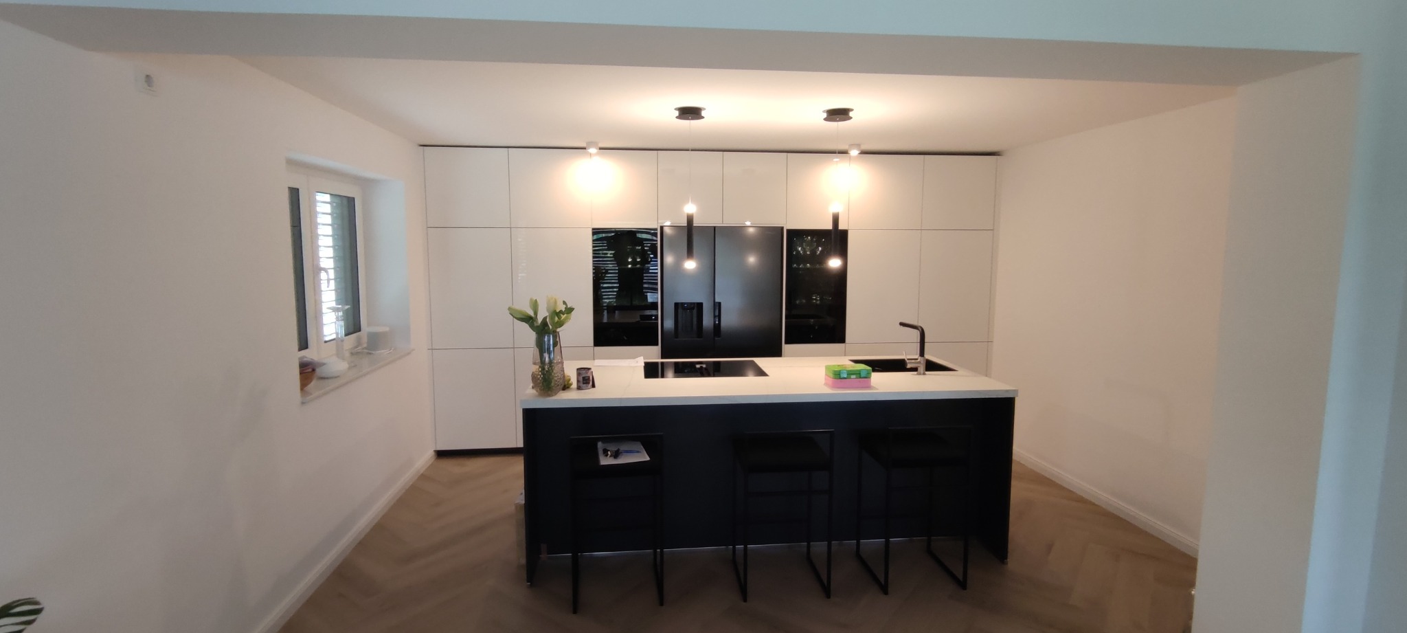 Nowoczesna kuchnia z białymi szafkami i czarną wyspą, oświetlona wiszącymi lampami, z drewnianą podłogą ułożoną w jodełkę.