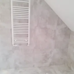 Zamontowany, biały grzejnik drabinkowy na ścianie z płytek w łazience na poddaszu, widoczne skosy.