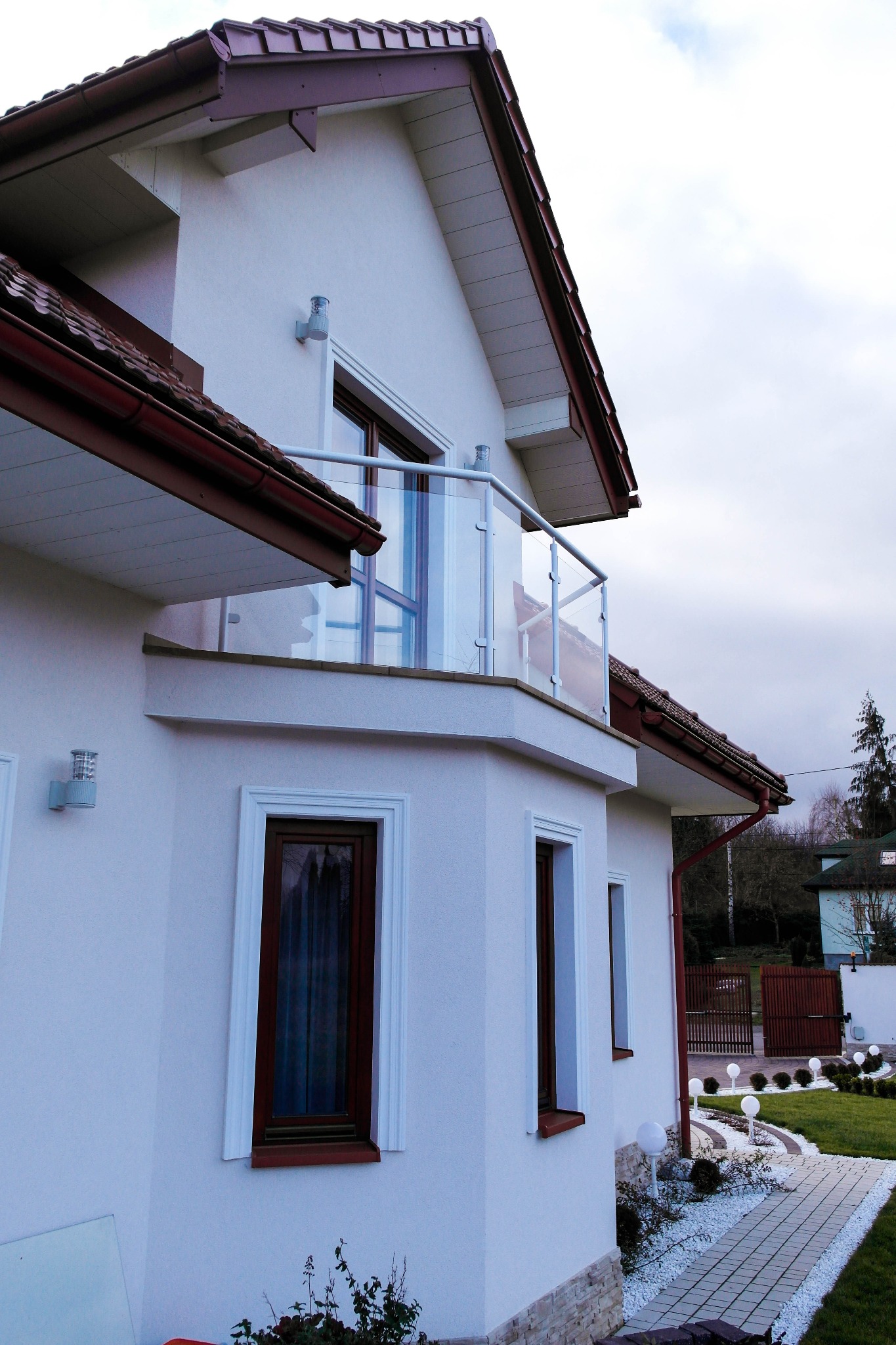 Biały dom z balkonem zabezpieczonym szklaną balustradą, widok z boku na elewację i fragment ogrodu z oświetleniem.