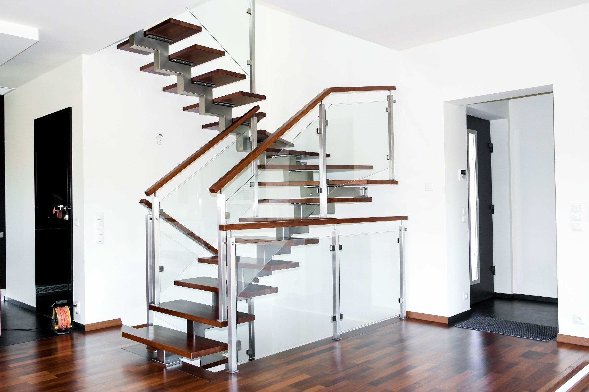 Nowoczesne schody z drewnianymi stopniami, metalowymi wspornikami i szklaną balustradą w jasnym wnętrzu z parkietem.