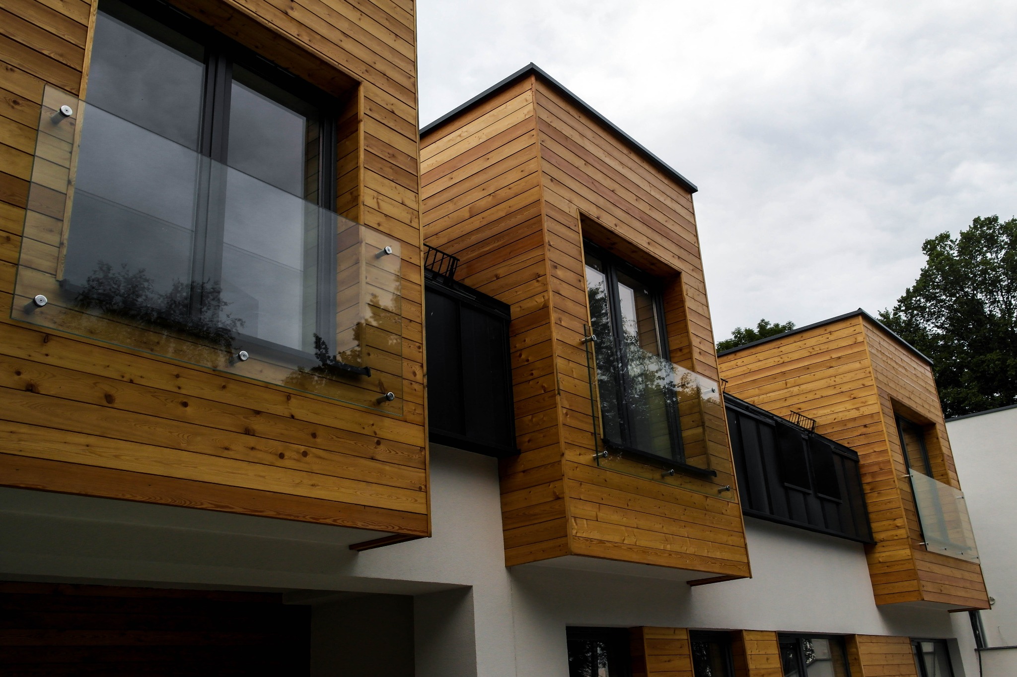 Nowoczesna elewacja budynku z drewnianą okładziną i szklanymi balustradami balkonowymi mocowanymi punktowo, widok z dołu na pochmurne niebo.