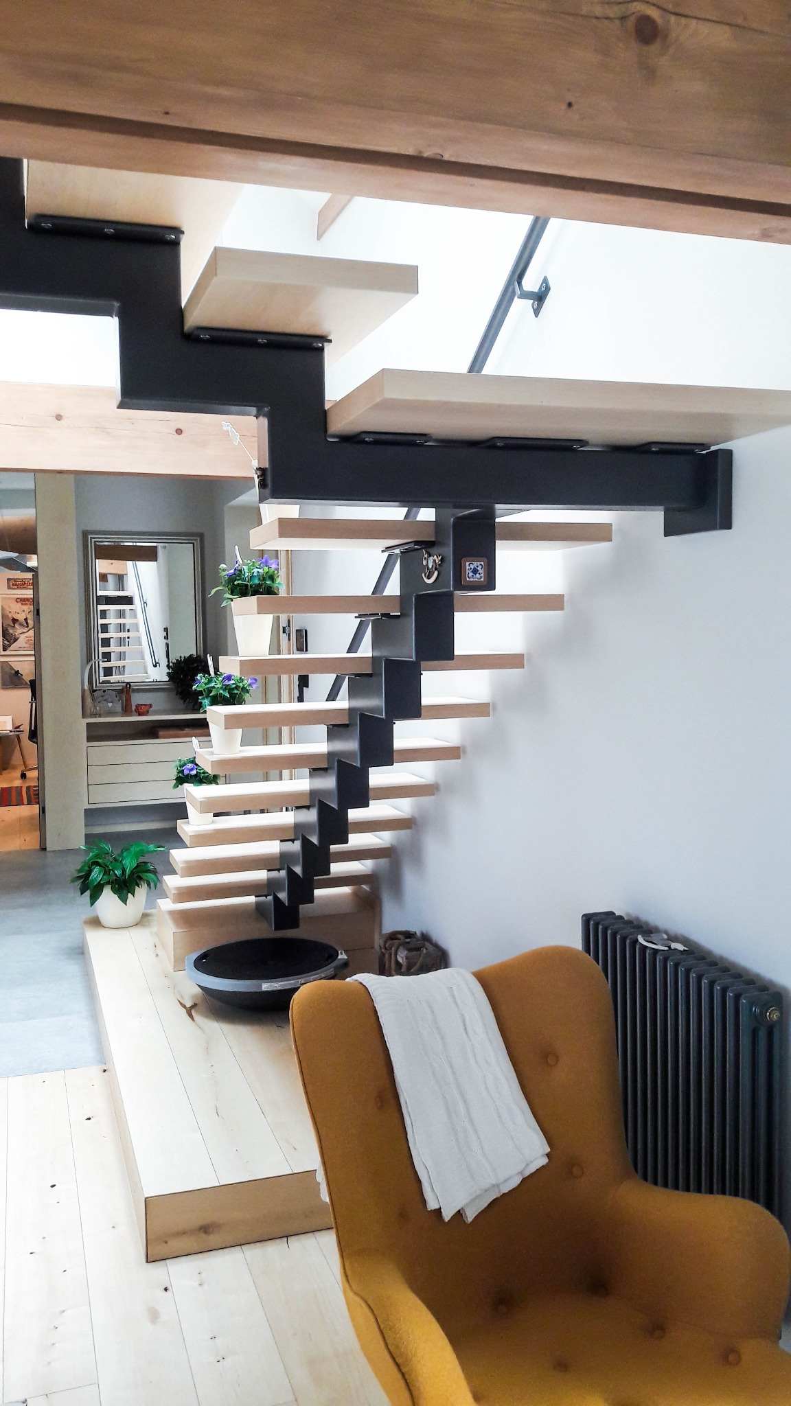 Nowoczesne schody drewniane z czarną, metalową konstrukcją nośną, w jasnym wnętrzu z drewnianą podłogą i musztardowym fotelem.