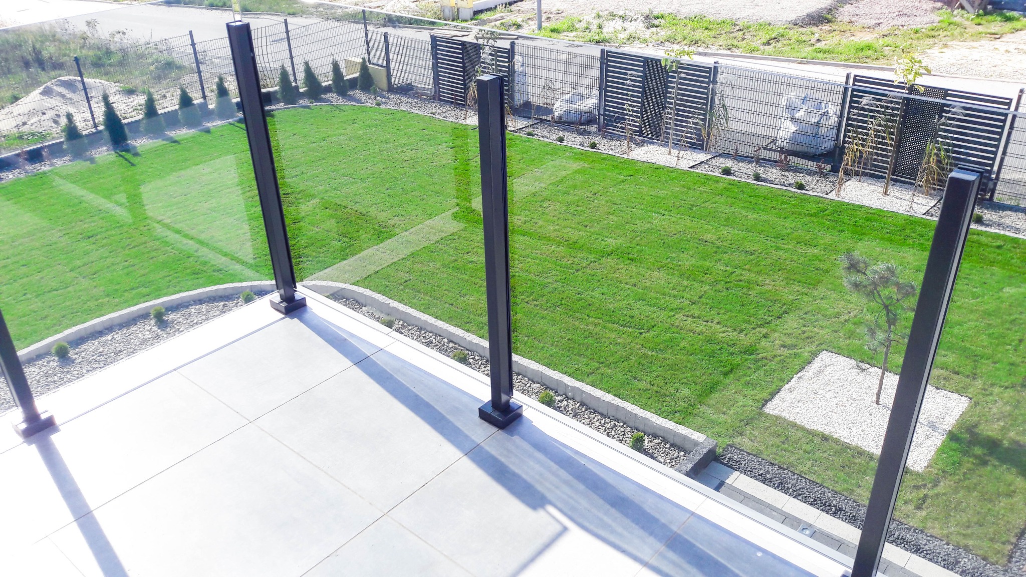 Nowoczesna balustrada szklana z czarnymi słupkami na tarasie, widok z góry na zadbany trawnik i ogrodzenie.