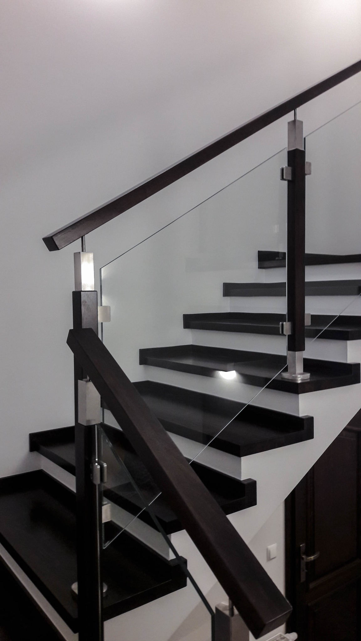 Nowoczesne schody z ciemnego drewna z podświetleniem stopni, zabezpieczone szklaną balustradą z elementami ze stali nierdzewnej.