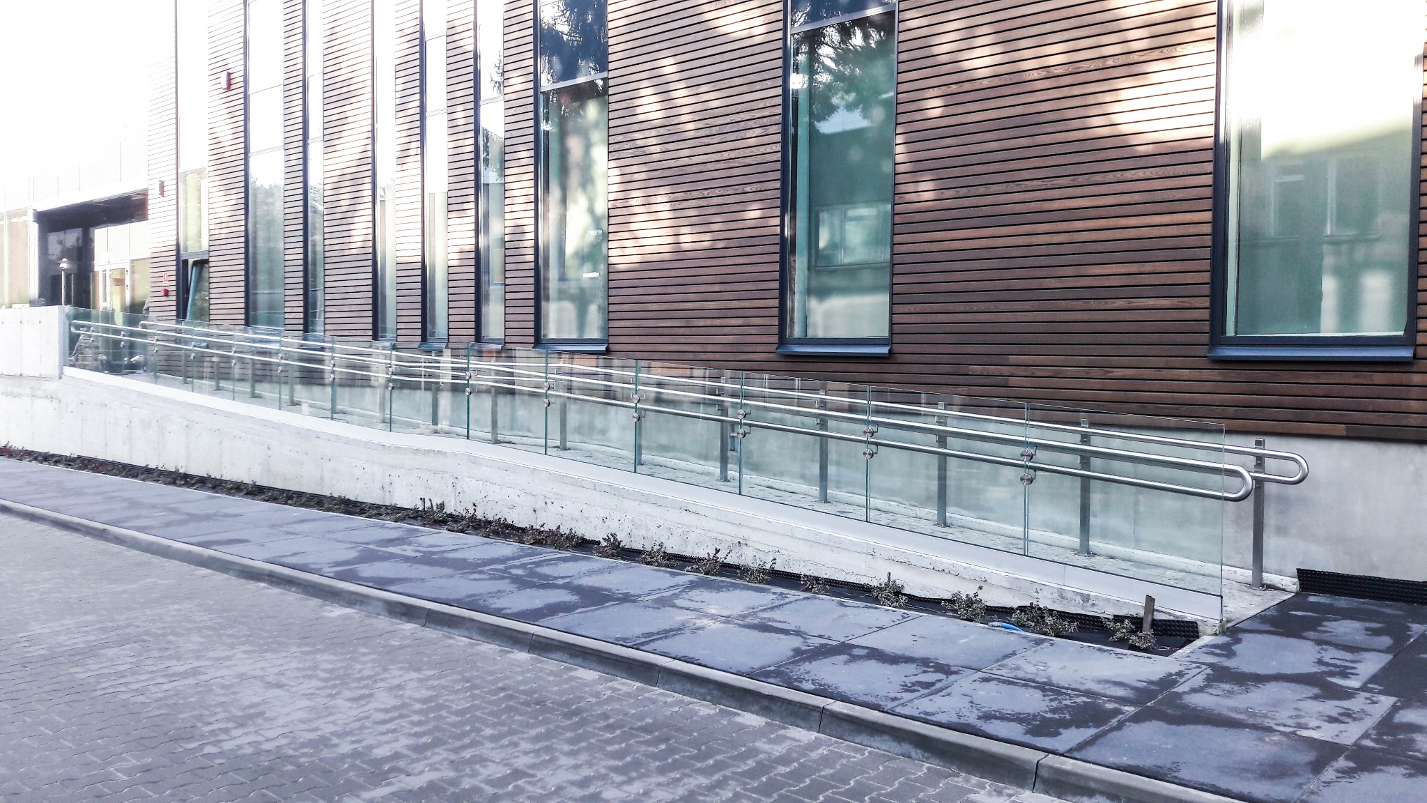 Szklana balustrada z metalowymi poręczami zabezpieczająca podjazd dla niepełnosprawnych w Krakowie, na tle budynku z drewnianą elewacją i dużymi oknami.