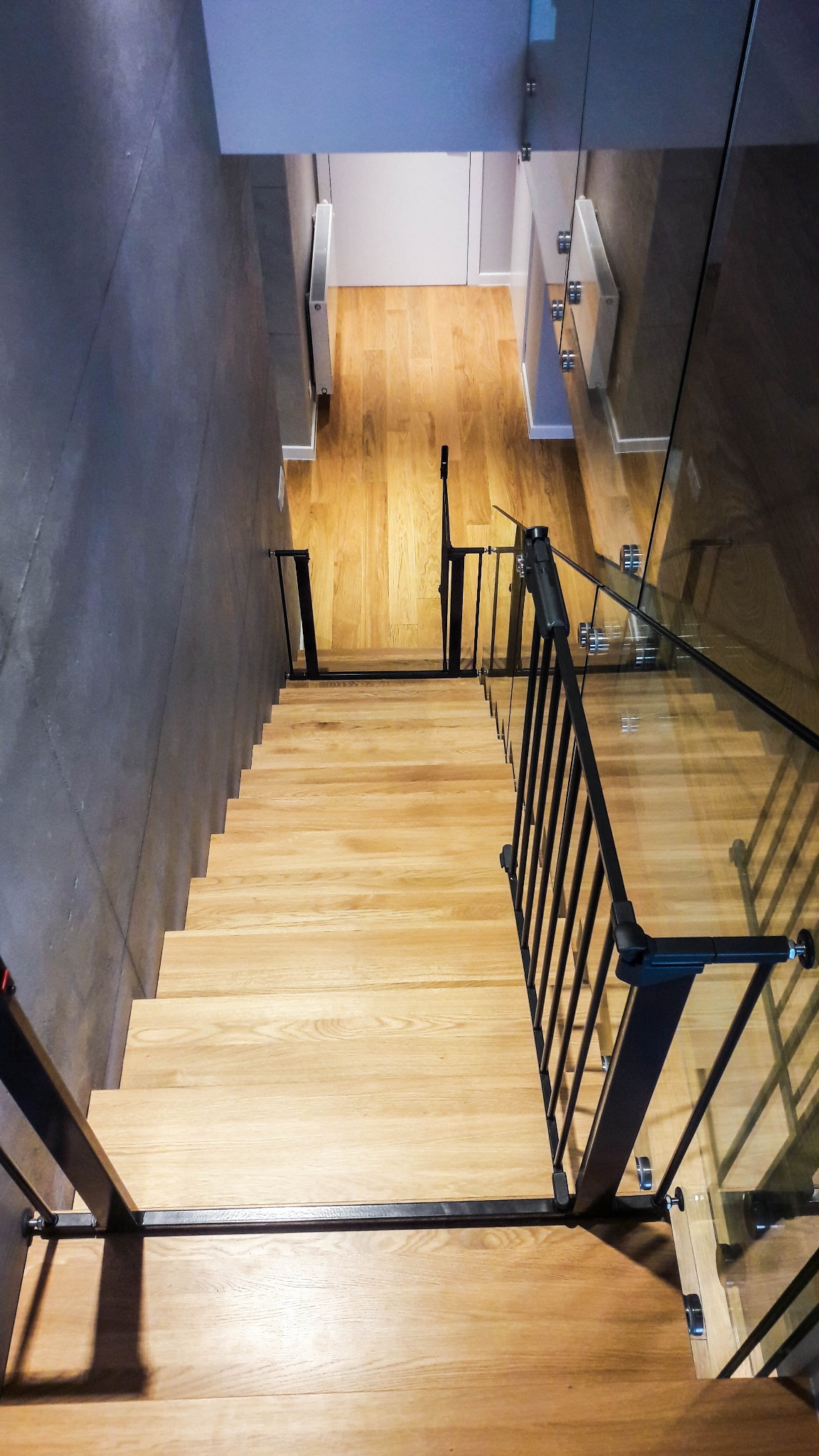 Nowoczesne, jasne schody z drewna dębowego, z czarną, metalową balustradą po jednej stronie i szklaną balustradą po drugiej, widok z góry.