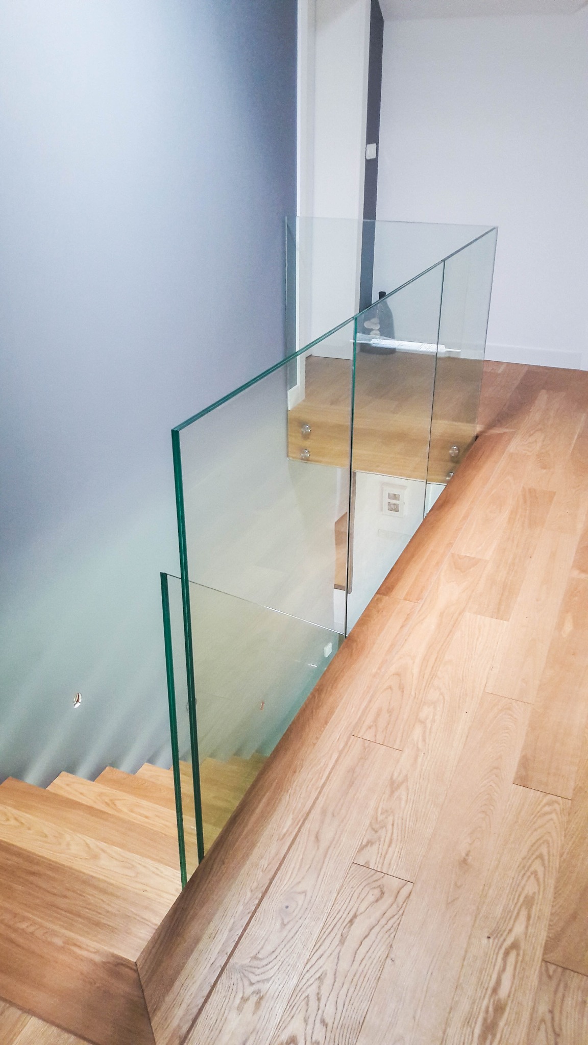 Nowoczesne schody drewniane z balustradą ze szkła hartowanego, mocowaną na metalowych uchwytach, widok z góry na stopnie i podłogę z jasnego drewna.