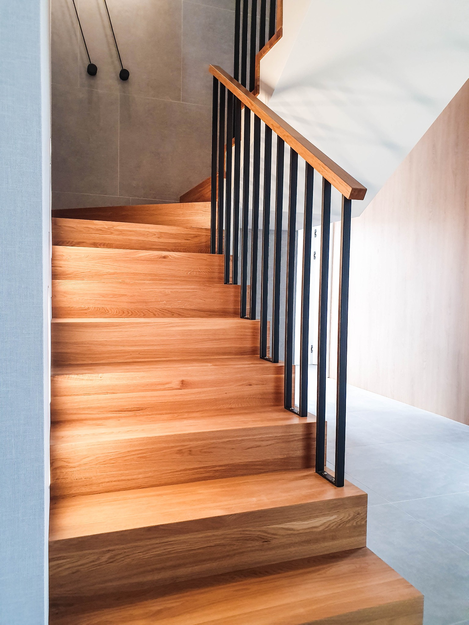 Nowoczesne schody z drewna o jasnym odcieniu, z czarnymi, pionowymi elementami balustrady i drewnianą poręczą, w minimalistycznym wnętrzu z szarymi płytkami podłogowymi i jasnymi ścianami.