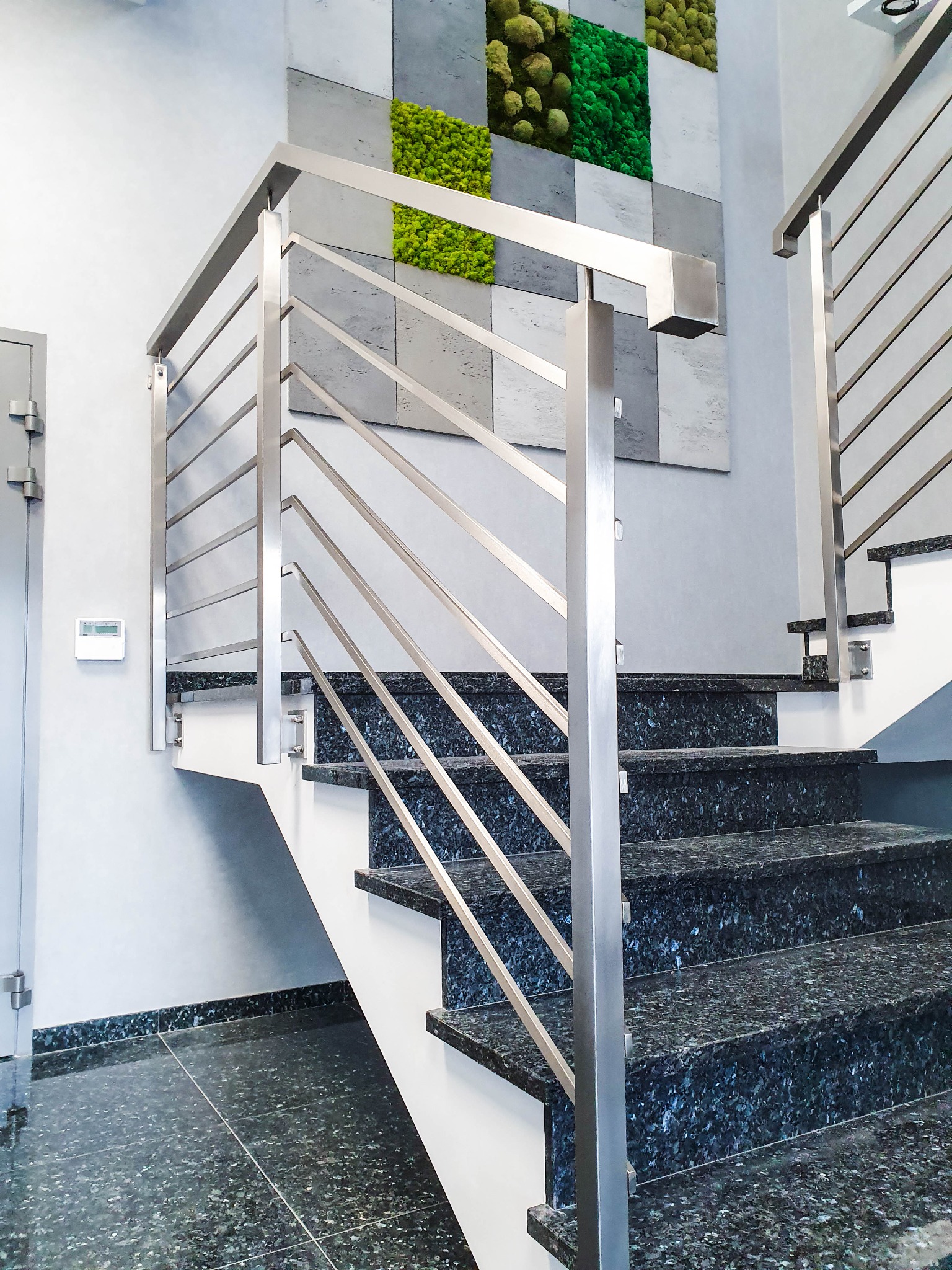Schody z ciemnego granitu z nowoczesną balustradą ze stali nierdzewnej, na ścianie dekoracja z mchu.