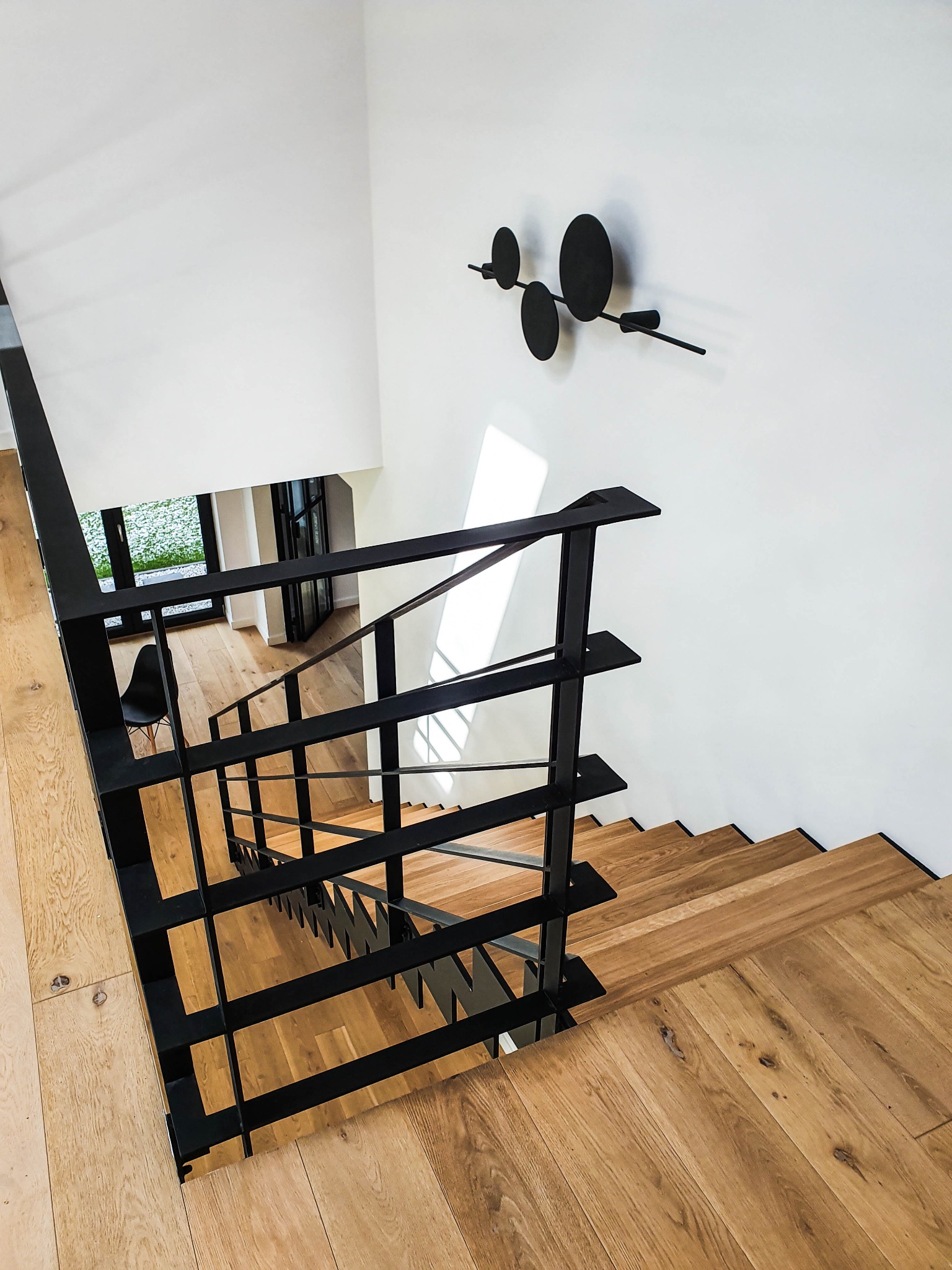 Nowoczesne schody z drewnianymi stopniami i czarną, geometryczną balustradą, widok z góry, minimalistyczny design, na ścianie dekoracja w kształcie czarnych kółek.