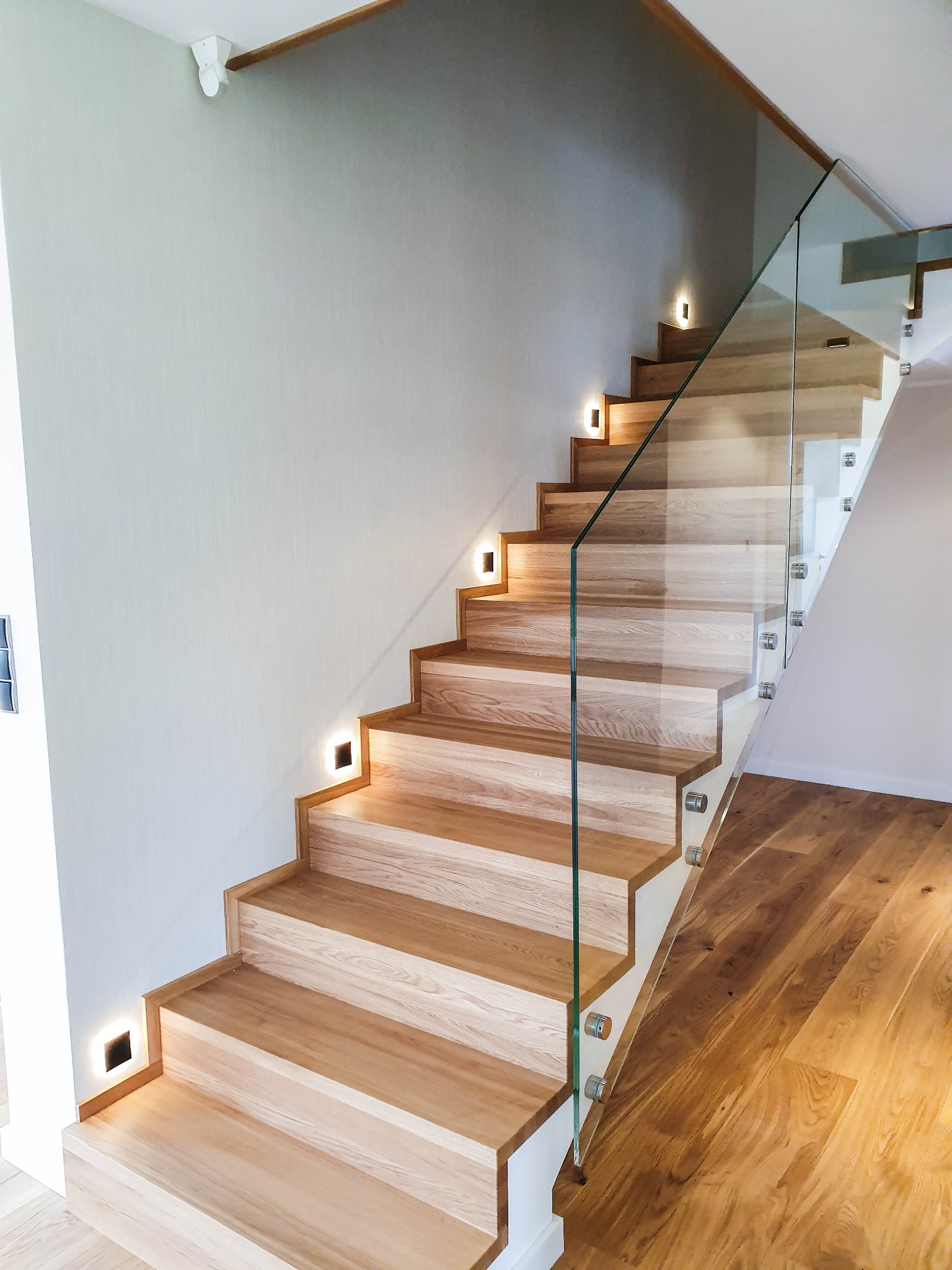 Nowoczesne schody drewniane z podświetleniem LED w stopniach i balustradą ze szkła mocowaną punktowo, widok z dołu na drewnianą podłogę