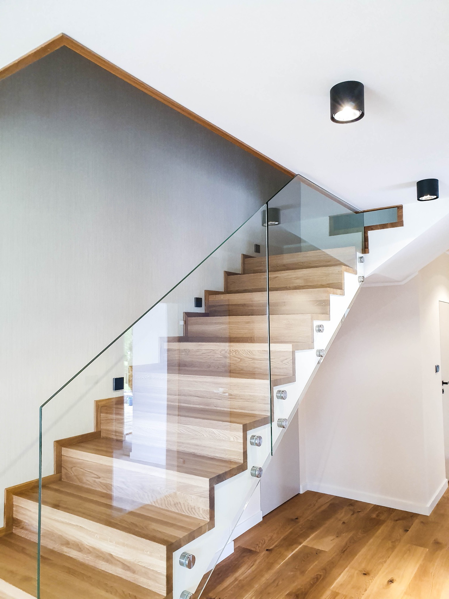 Nowoczesne schody z drewna z przezroczystą balustradą szklaną, mocowaną za pomocą metalowych uchwytów punktowych, oświetlone czarnymi lampami sufitowymi, na tle szarej ściany z drewnianym...
