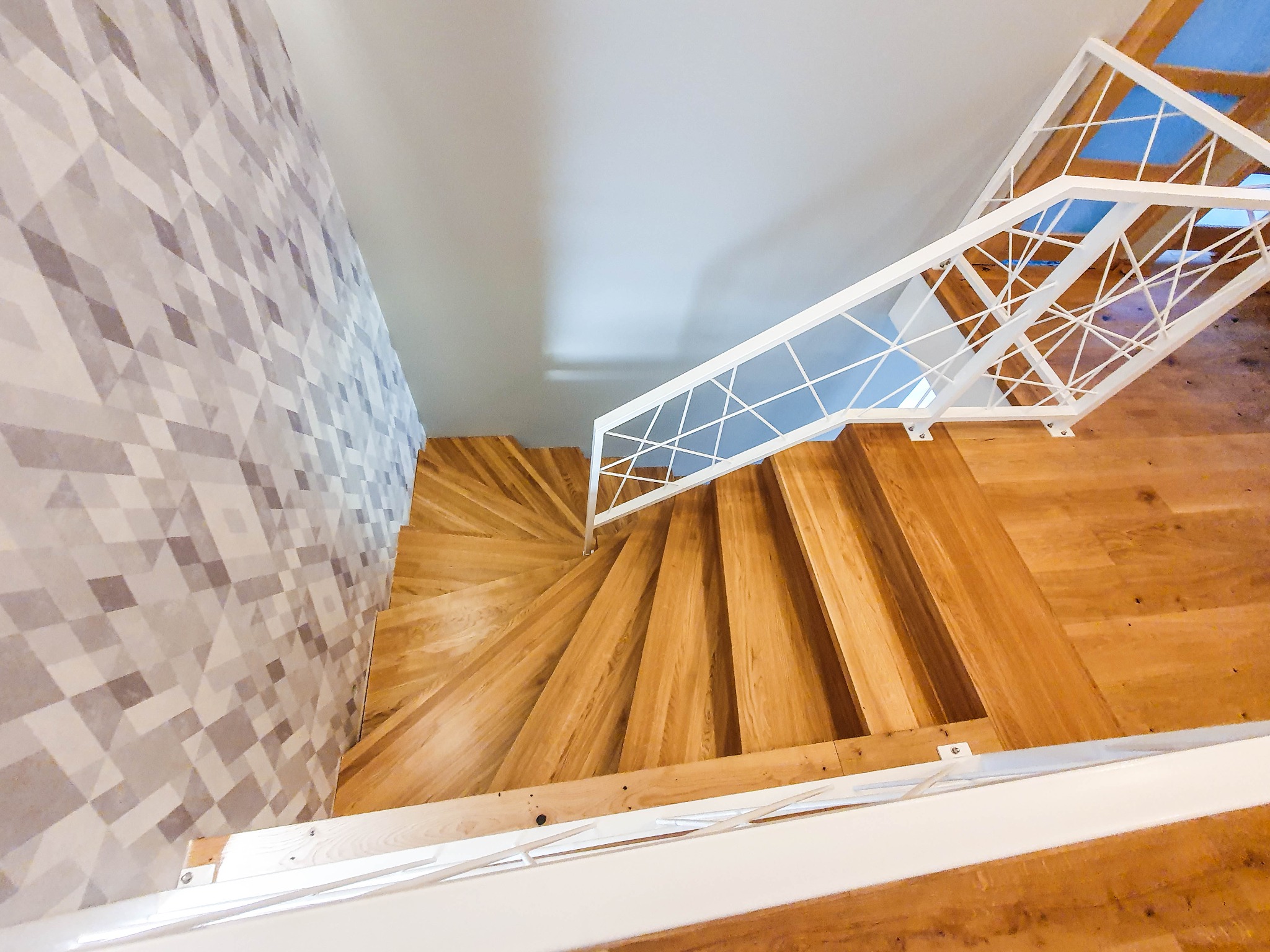 Spiralne schody z jasnego drewna, biała, geometryczna balustrada, widok z góry, wzorzysta ściana w tle.