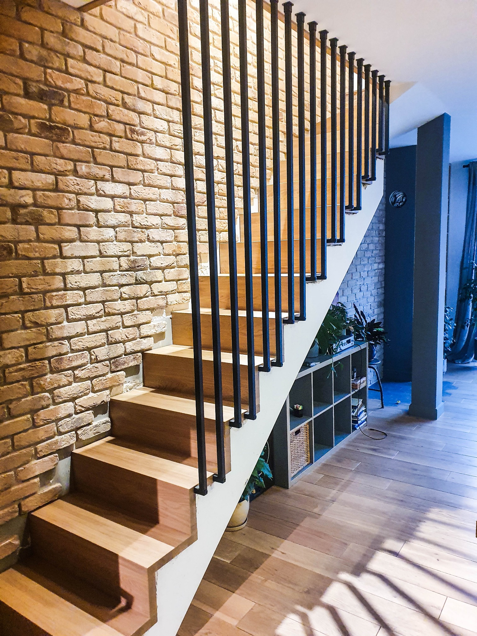 Nowoczesne schody z drewnianymi stopniami i czarnymi, pionowymi balustradami na tle ceglanej ściany, z widoczną półką z roślinami pod schodami oraz drewnianą podłogą.