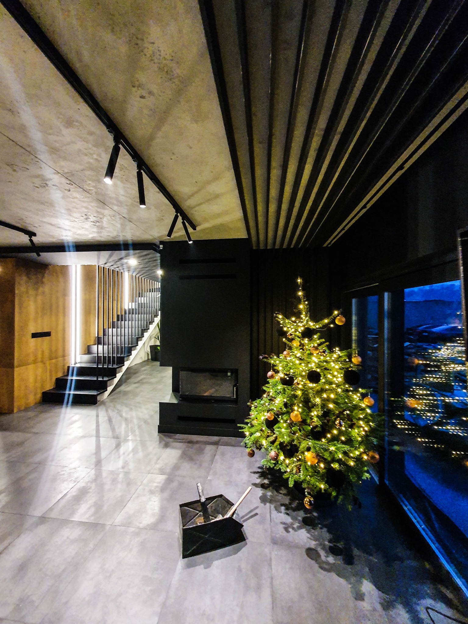 Nowoczesne wnętrze domu z czarnymi schodami z metalową balustradą, kominkiem i choinką, widok na nocne miasto za oknem.