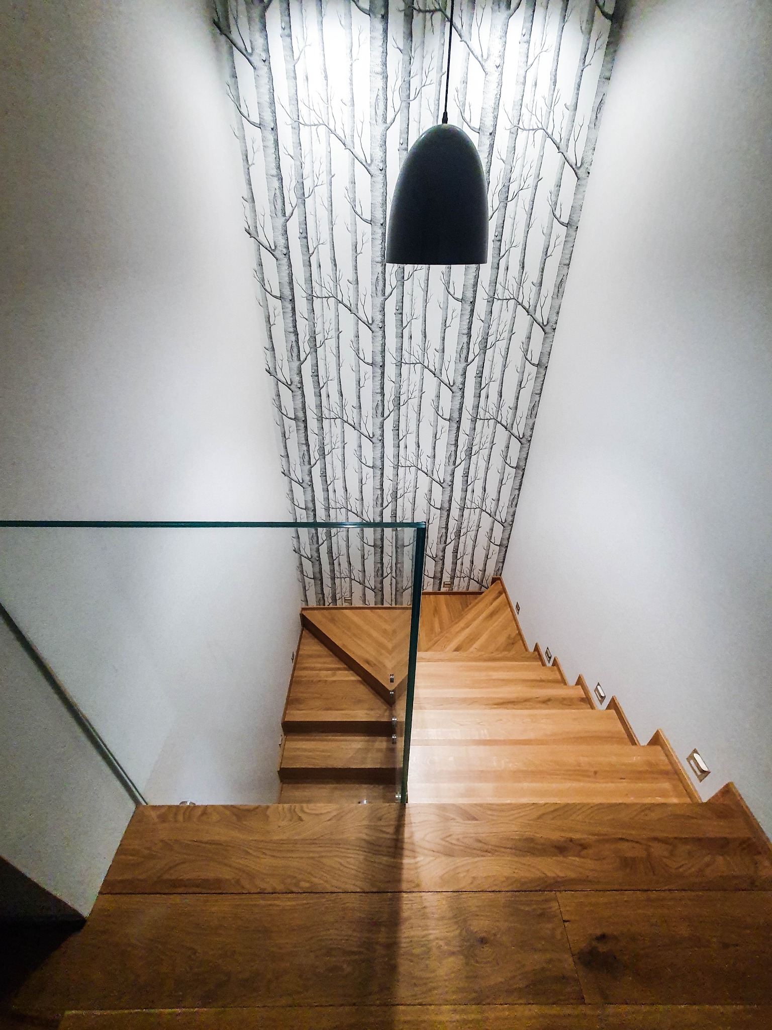 Widok z góry na dębowe schody z podświetleniem i szklaną balustradą, ściana z tapetą w motywie brzozowego lasu, czarna lampa wisząca.