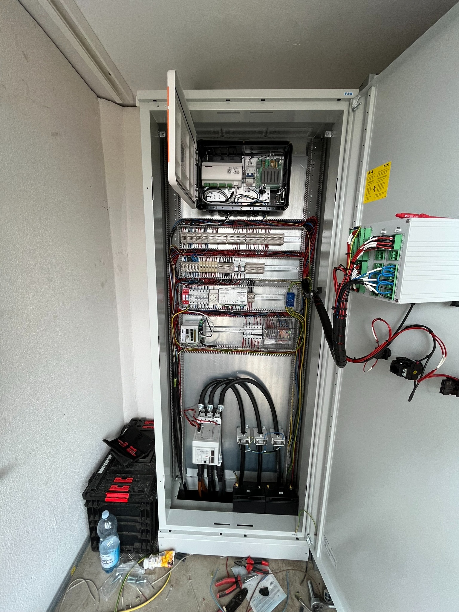 Otwarta szafa elektryczna z widocznymi kablami, bezpiecznikami i elementami sterującymi. Na podłodze narzędzia, butelka wody i resztki instalacji.